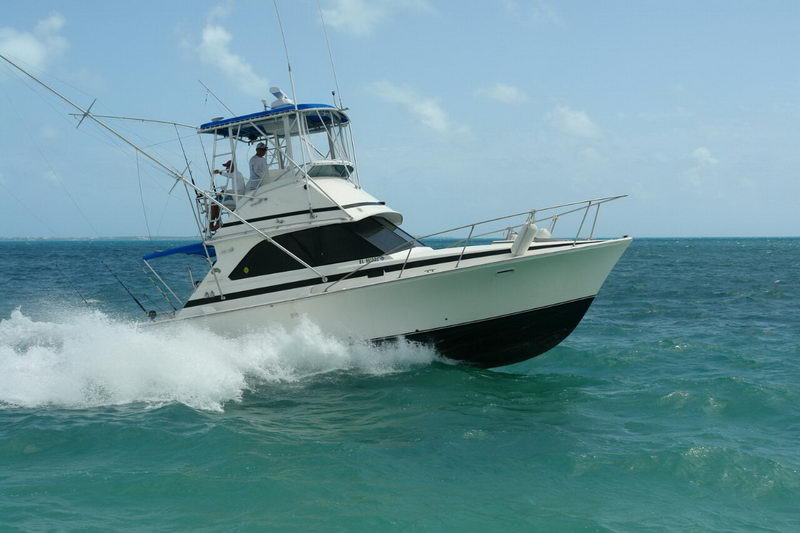 White sportfishing convertible with a flybridge turning at speed across turquoise sea, throwing a bow wave. Two anglers stand on the flybridge and outriggers extend to the side under a hazy sky.