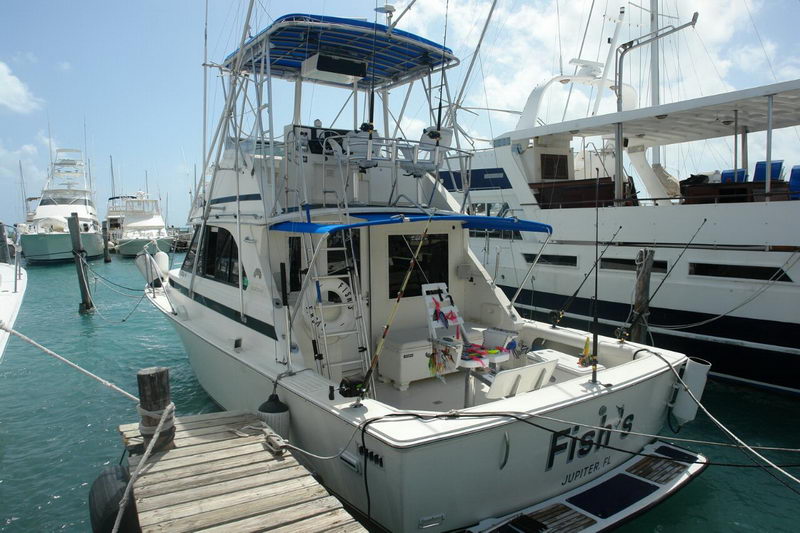 Stern view of a sportfishing boat named 'Fish' from Jupiter FL docked stern-to at a marina, with fishing rods, tackle and a cooler in the cockpit. Other boats and pilings sit in turquoise water around it.