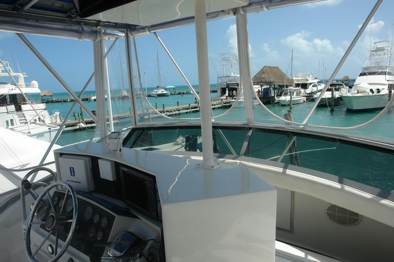 View from the flybridge helm of a sportfishing boat, showing the wheel, Furuno electronics and gauges, looking out over a marina with moored boats, docks and a thatched palapa across turquoise water.