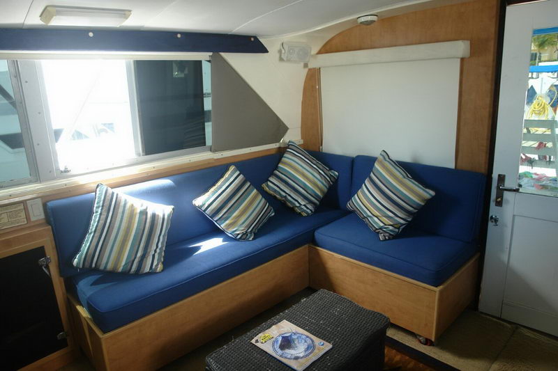 Interior salon of a sportfishing boat with an L-shaped settee upholstered in royal blue, several striped throw pillows, light wood paneling, a side window and a wood-and-glass door.