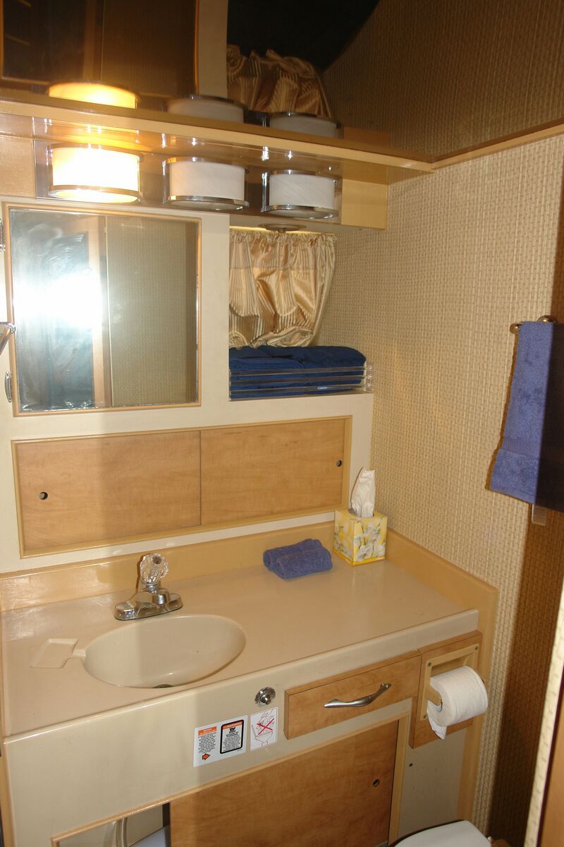 Interior head of a sportfishing boat with a cream vanity and oval sink, a mirrored cabinet, folded blue towels, a tissue box and toilet paper, surrounded by woven beige wall paneling.