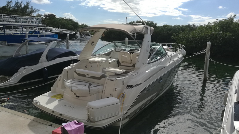Silver-hull Monterey cruiser with a tan bimini moored bow-out at a dock, its tan cockpit seating visible. Mangroves and other boats sit behind under a partly cloudy sky.