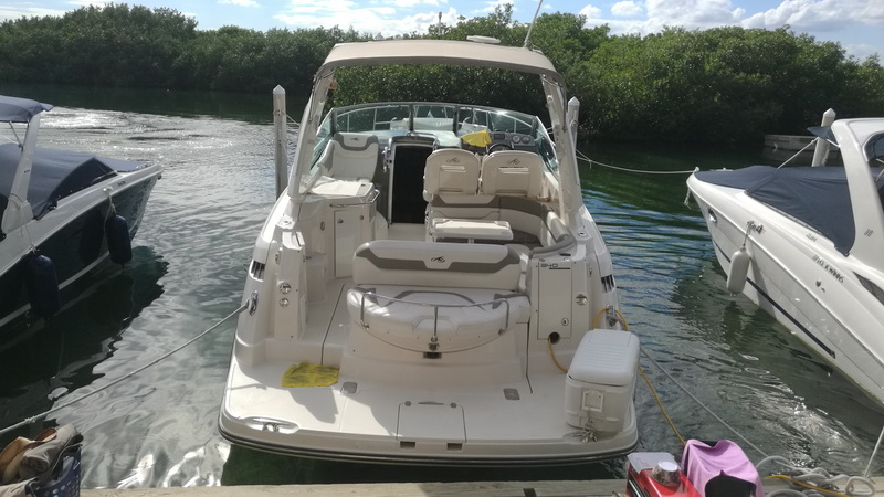 Front view of a silver-hull Monterey cruiser with a tan bimini moored between two other boats, showing the cockpit seating and windshield. Mangroves and green water surround it.