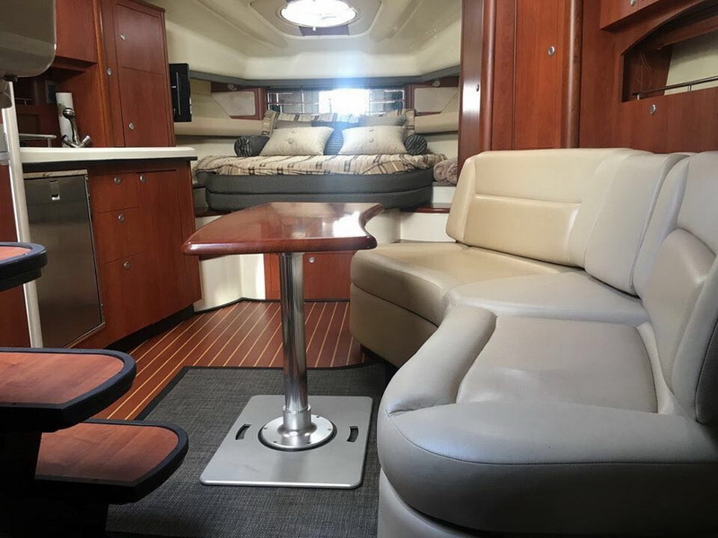 Interior cabin of a cruiser with a curved grey leather settee, a cherry wood dinette table, a mid-cabin berth with patterned pillows behind, a galley at left and teak-and-holly flooring.