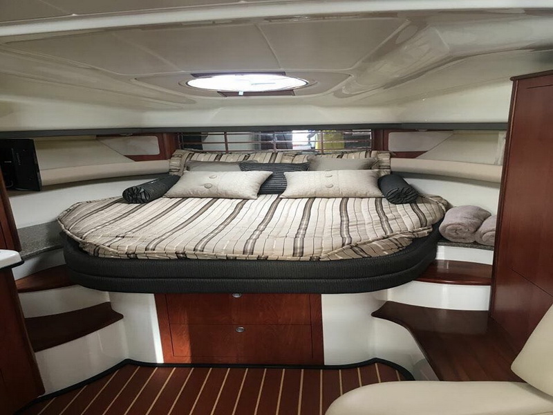 Mid-cabin double berth of a cruiser with a striped beige bedspread and matching pillows, glossy cherry wood cabinets on each side and a round overhead hatch letting in light.