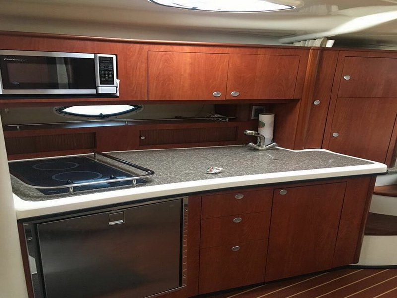 Galley of a cruiser with glossy cherry wood cabinets, a stainless microwave, a two-burner cooktop, a stainless fridge, a sink and a beige countertop, with a porthole above.