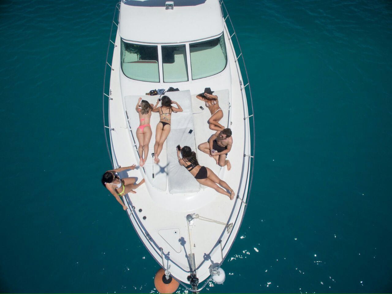Toma con dron mirando hacia abajo a la proa del Sea Ray 44 con un grupo de amigas en traje de baño tomando sol sobre el agua azul profundo