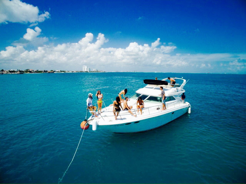 Yate blanco Sea Ray 44 con flybridge anclado en aguas turquesas con invitados en la proa y el skyline de Cancún al fondo