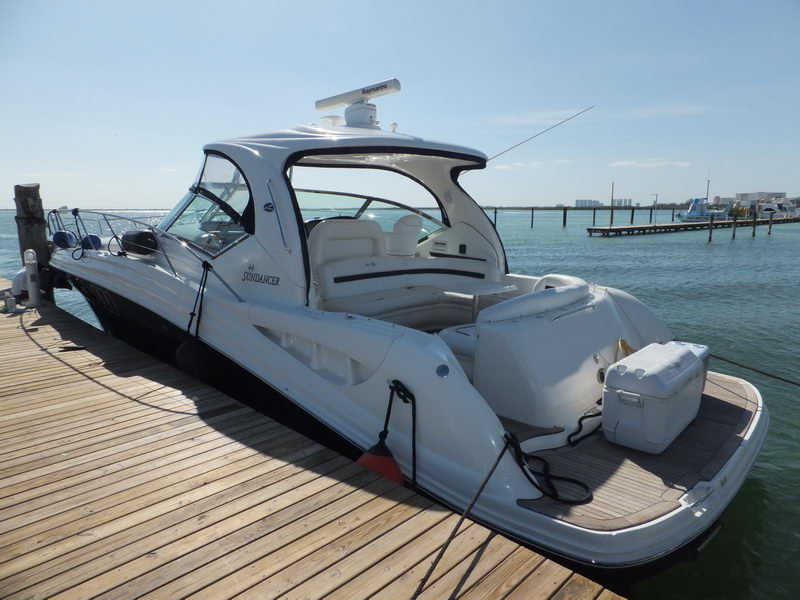 Stern quarter of a white Sea Ray Sundancer tied alongside a wooden dock, showing the radar arch, a cooler on the swim platform and turquoise water with distant piers behind.