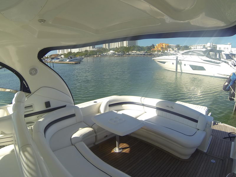 Cockpit lounge of a Sea Ray Sundancer with white seating and a low table, looking out across calm marina water toward another moored yacht and waterfront buildings in bright sun.