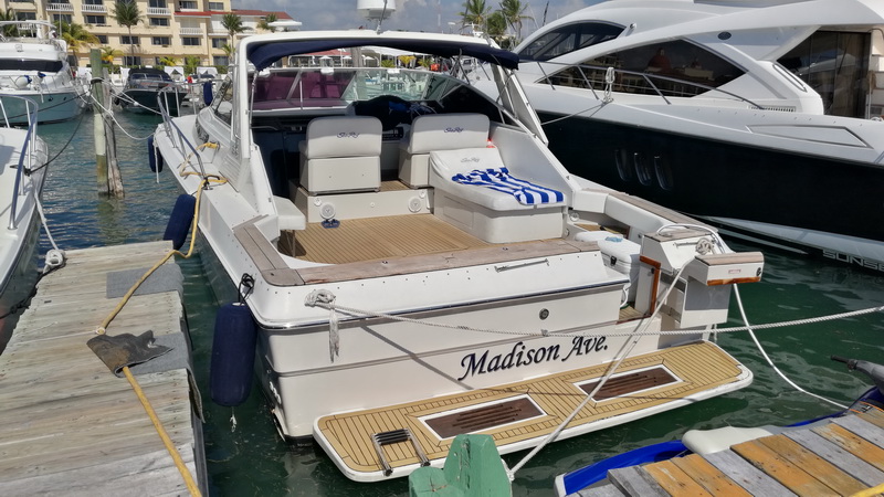 Stern view of a Sea Ray express cruiser named 'Madison Ave.' at a dock, with a teak swim platform, a blue-and-white striped cockpit cushion, and adjacent boats including a larger Sunseeker. Sunny marina.