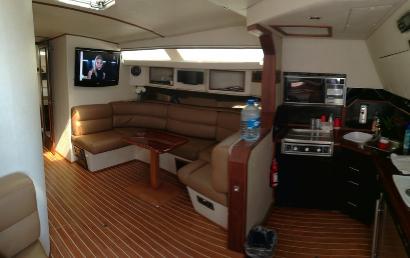 Interior salon with a tan and brown leather L-shaped sofa, a wood dinette table, a flat-screen TV, a galley with stove and microwave, and a teak-look sole. Bright cabin.