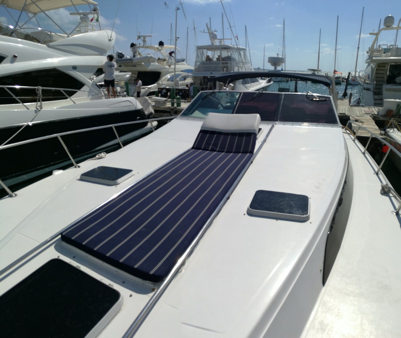 Foredeck of the express cruiser with a navy pinstripe sunpad cushion and a white bolster pillow on the bow, looking back toward a crowded marina full of yachts and masts in bright sun.