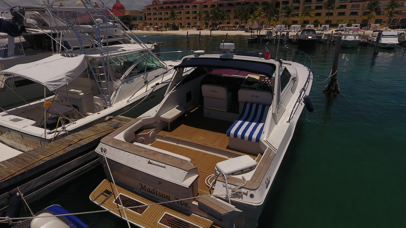 Elevated view of the express cruiser 'Madison Ave.' tied at a dock, cockpit fitted with a blue-and-white striped sun lounger, with a beach resort and palms across the marina behind.
