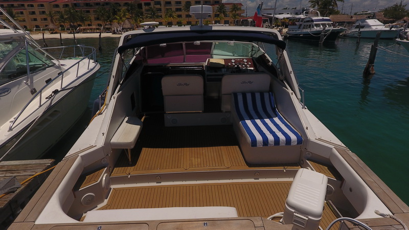 Aft cockpit of the Sea Ray seen from above, with white seats, a blue-and-white striped sun lounger cushion and teak sole, the marina and a beachfront resort visible beyond.