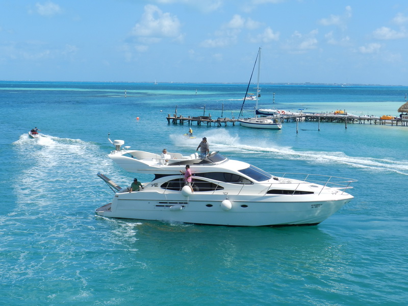 White Azimut-style flybridge motor yacht underway on bright turquoise water near a long pier, with people aboard, a jet ski and a moored sailboat nearby; Cancun shoreline in the distance.