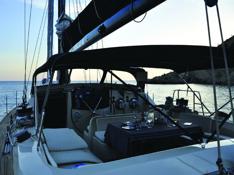 Cockpit of a sailing yacht at dusk: twin helm wheels, polished winches, cushioned seating and a table set with glasses and candles, rocky coastline and calm sea in soft evening light.
