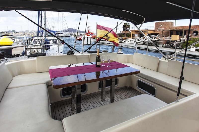 Cockpit dining setup with cream bench seating around a wood table with a burgundy runner, champagne bottle and two glasses, a Spanish flag flying, marina and harbor buildings behind.