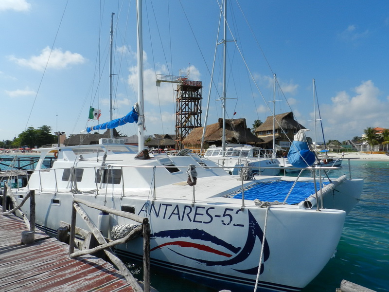 White sailing catamaran 'Antares 52' moored at a wooden dock with a Mexican flag, beach palapas, a wooden lookout tower and other catamarans behind, on clear turquoise water under a blue sky.