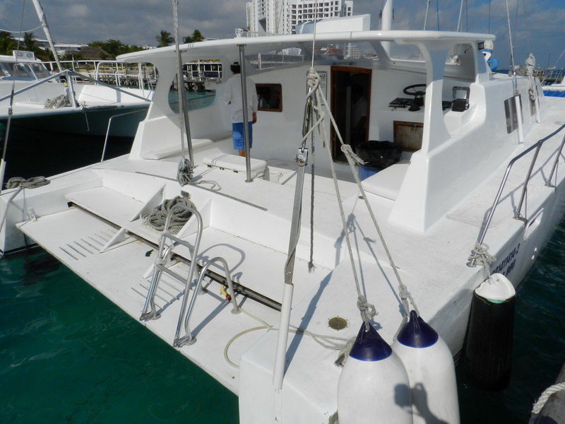 Stern view of a white sailing catamaran at a dock with a crew member aboard, blue fenders hanging, other catamarans and the marina behind, on turquoise water under a partly cloudy sky.