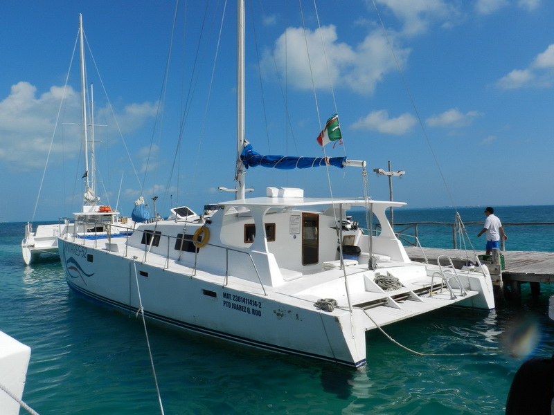 White sailing catamaran moored at a dock with a furled mainsail and a Mexican flag, a crew member standing at right, another catamaran alongside, on clear turquoise water under a blue sky with clouds.