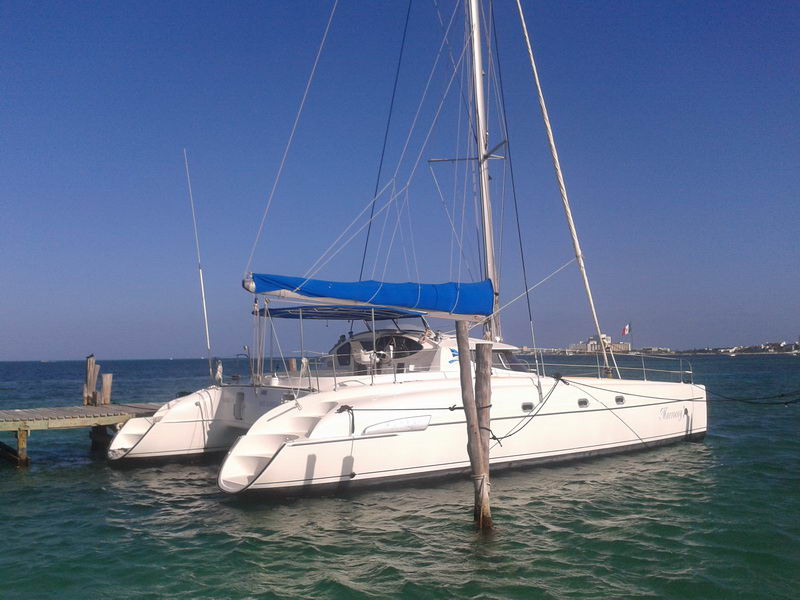 White cruising catamaran 'Morrocoy' with a blue sail cover moored at a dock in turquoise water, mast raised, shoreline and buildings behind on a sunny day.
