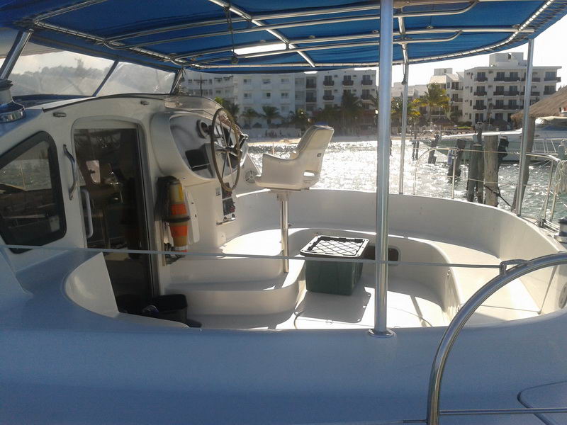 Cockpit and helm of the 'Morrocoy' catamaran under a blue Bimini, showing the steering wheel, a raised helm seat and cushioned seating, shore with buildings visible.