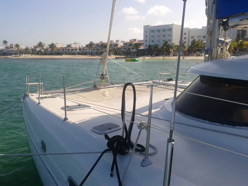 Foredeck of the 'Morrocoy' catamaran with anchor lines, looking toward a beach with hotels and palm trees over turquoise water, sunny.