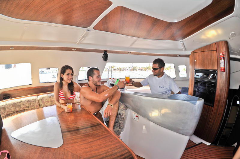 Interior salon of a catamaran with three people (a woman in a red bikini, a man and a crew member) at a wood-finished bar/galley with bright wood paneling, drinks being served.