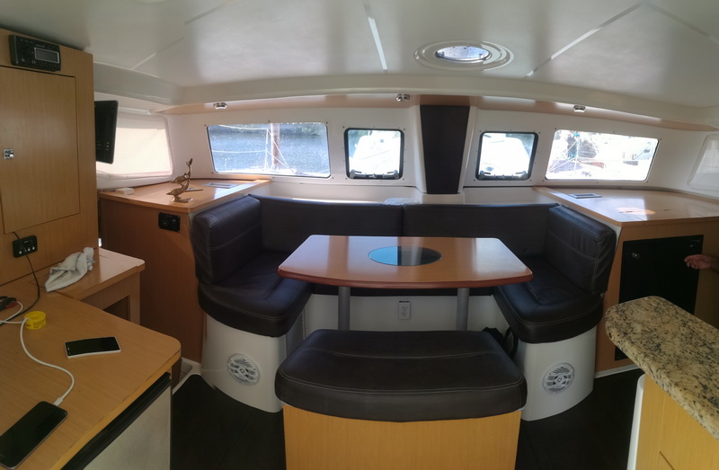Interior salon of a catamaran with a dark leather U-shaped settee, a wood table with a circular inset, light wood cabinetry and windows, a phone charging on the counter.