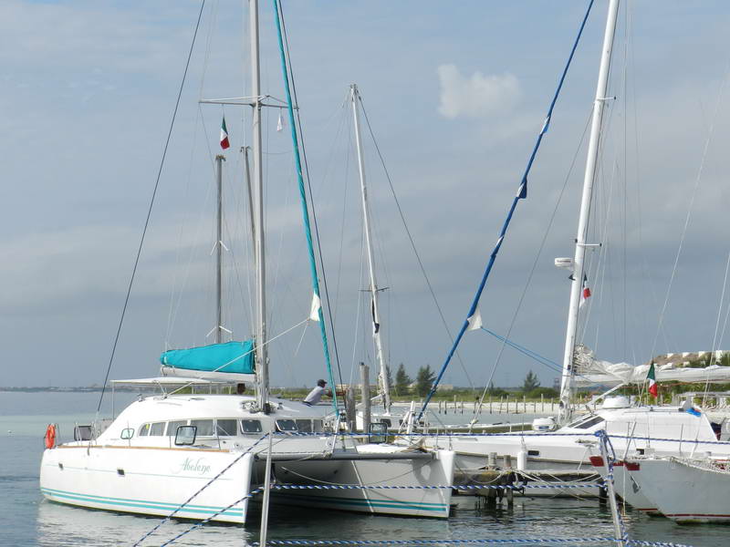 White sailing catamaran named 'Abelaye' moored at a marina alongside several other sailboats with masts and rigging, teal sail cover, Mexican flag on the mast, a sandy point and cloudy sky behind.