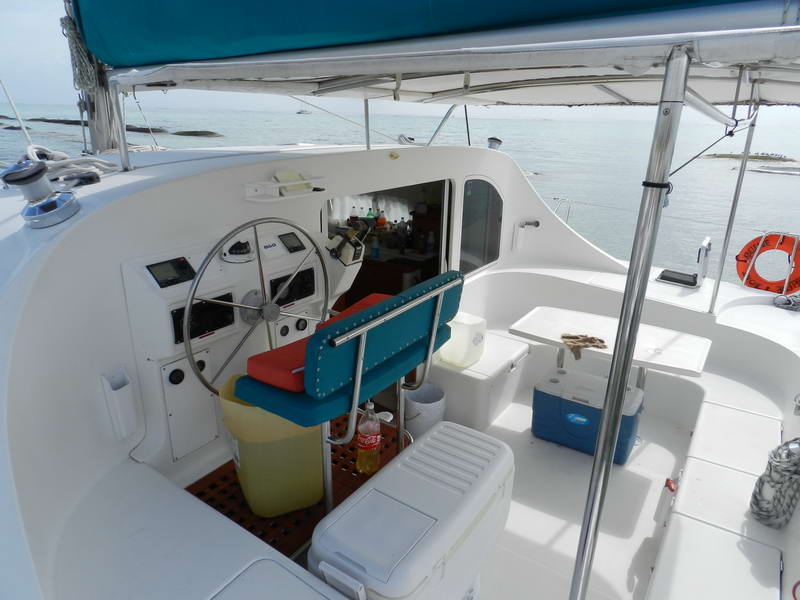 Cockpit and helm station of a sailing catamaran showing the stainless steering wheel, instrument panel, a red-and-teal helm seat, several coolers and supplies under a bimini, looking out over shallow turquoise reef water.