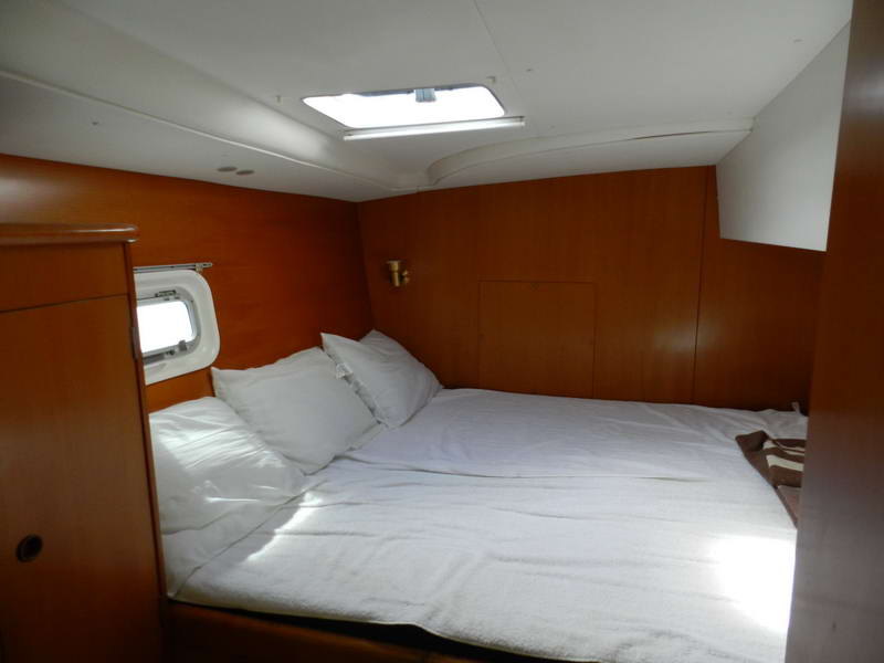 Interior sleeping cabin of a catamaran with a double bed made up in white linens, warm wood paneling, an overhead hatch letting in daylight, a small porthole and a brass reading light.