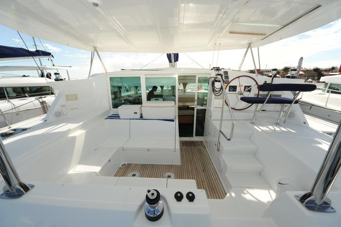 Aft cockpit of a Lagoon sailing catamaran at a marina showing the helm wheel, a cushioned bench, teak steps leading up and sliding glass doors into the salon, with other catamarans alongside.