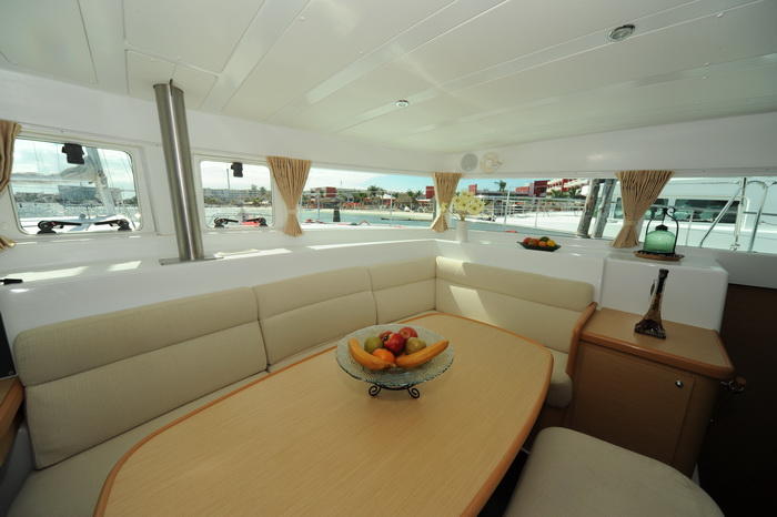 Interior salon of a Lagoon catamaran with a cream U-shaped settee, an oval wood dining table holding a bowl of fruit, panoramic curtained windows, light wood cabinetry and a marina view outside.