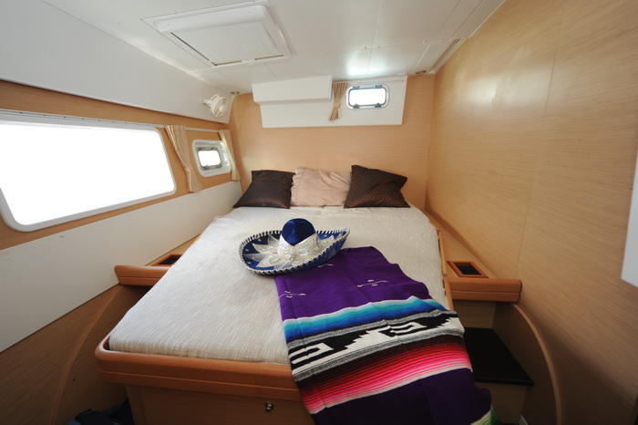Interior double cabin of a catamaran with a bed dressed in white and topped with a blue Mexican sombrero and a colorful striped serape blanket, light wood paneling and a hull window.