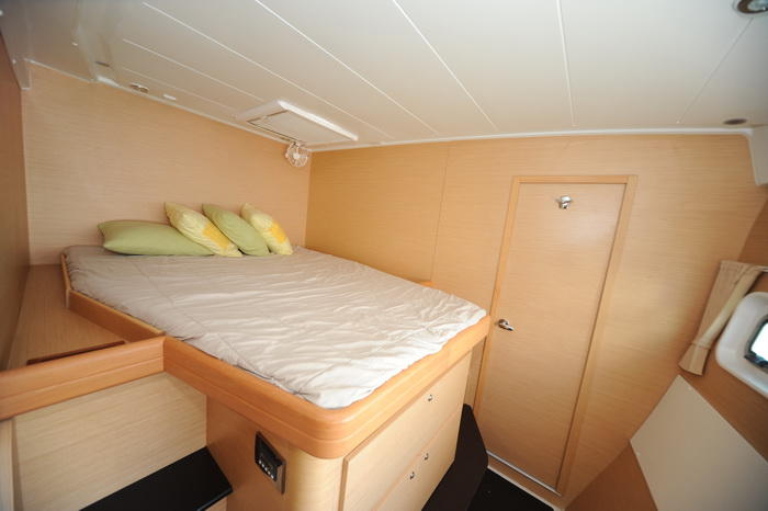 Interior double cabin of a catamaran with a bed made up in beige with green and yellow pillows, light wood paneling, a closed wooden door and a storage cabinet with a combination lock.