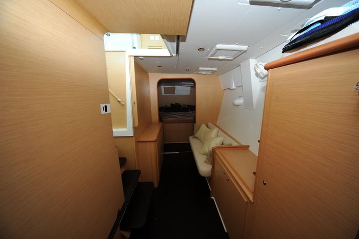 Interior of a catamaran hull showing a narrow passage with a small cream settee and pillows, light wood paneling, a glimpse of a berth ahead and storage units along a dark floor.