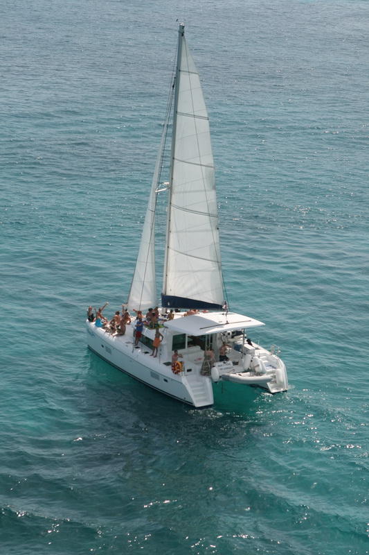 Aerial view of a white Lagoon sailing catamaran under sail with a group of passengers waving on the foredeck, sailing over clear turquoise Caribbean water on a sunny day.