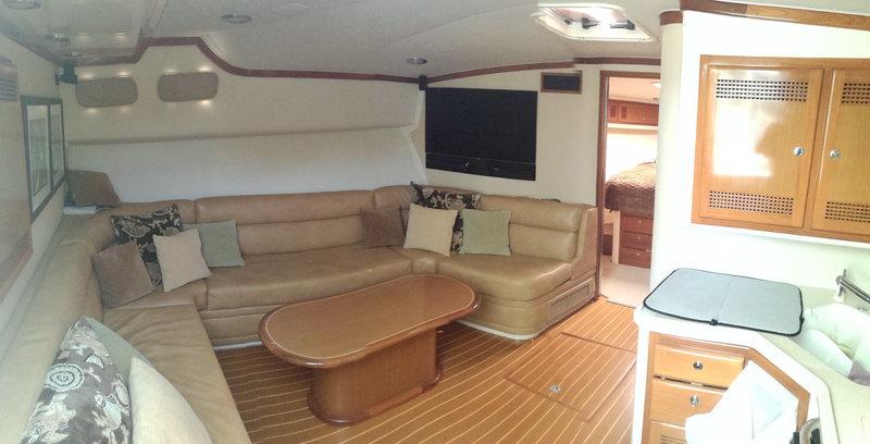 Interior salon of a sportfishing yacht with a tan leather sectional sofa, scatter cushions, a wall-mounted flat TV, teak-and-holly floor and a glimpse of a berth and galley cabinetry beyond.