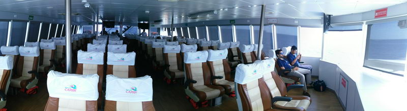 Interior passenger cabin of a large catamaran ferry (Barcos Caribe) with rows of beige and brown reclining seats fitted with white logo headrest covers; two passengers seated at the rear by the windows.