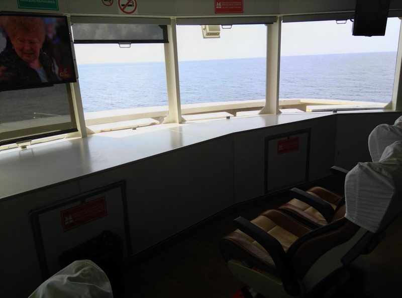 Interior of a passenger ferry showing the captain's-area front cabin: a long white console runs beneath panoramic windows that look out over flat open grey-blue sea. Reclining brown-and-cream leather seats sit at right and a small wall-mounted TV screen is at left.