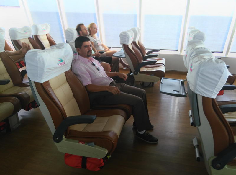 Inside a passenger ferry cabin with rows of brown-and-cream reclining leather seats topped by white logoed headrest covers. A man in a pink shirt sits in the foreground, other passengers behind, and large windows show bright ocean light. Wood-look flooring.