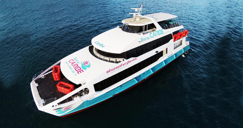 Exterior aerial view of a white Barcos Caribe express ferry catamaran underway on deep blue water. The hull carries turquoise trim and '#ExpressFerryService' branding, with red life rafts and an open foredeck visible from above.
