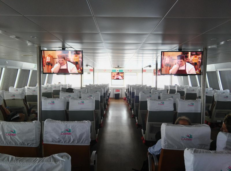 Economy interior cabin of a ferry seen down the central aisle: rows of grey seats with white logoed headrest covers on both sides, two flat TV screens mounted near the ceiling at front, and a few passengers seated. Bright windows along the sides.