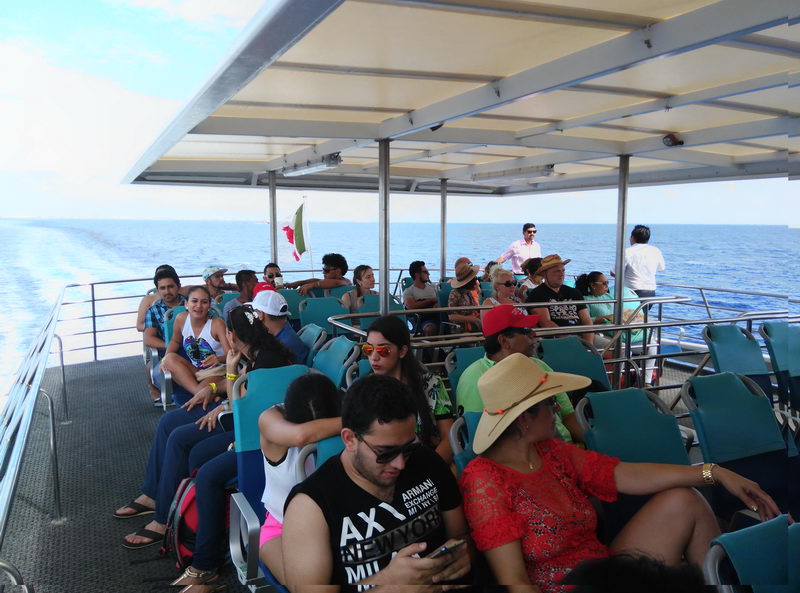 Open-air upper sun deck of a ferry crowded with seated passengers in turquoise plastic chairs under a white canopy roof. Bright sunny day, open ocean to the sides and a small Mexican flag flying. People in summer clothing and hats.