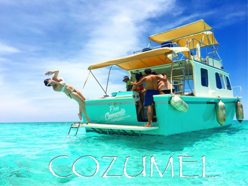 The turquoise 'Don Clemente' tour boat anchored on vivid turquoise water with a woman diving off the stern and a group of people gathered at the back. Yellow canopies shade the deck; a large 'COZUMEL' text overlay sits across the bottom.