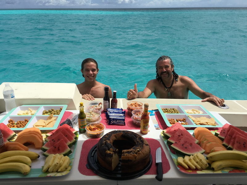 Two smiling men stand in turquoise water behind a boat's white swim platform that is laid out with snack platters of watermelon, papaya, banana, olives, chips, salsa and a bundt cake. One gives a thumbs-up; open sea behind.