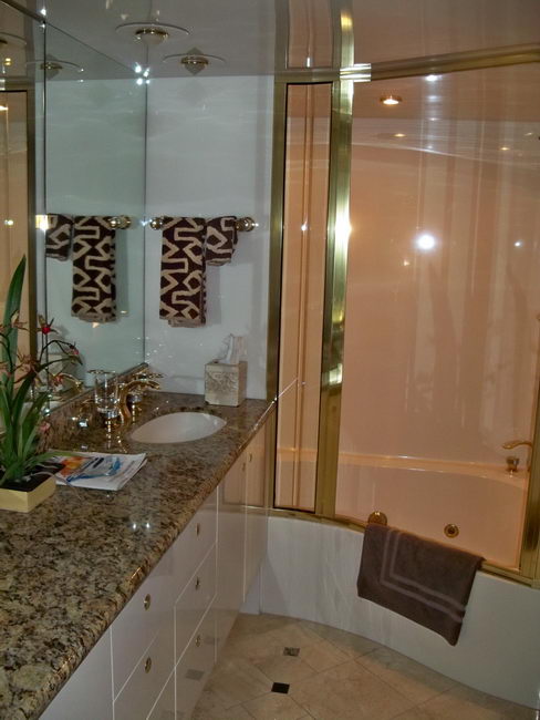 Yacht interior bathroom with a beige granite vanity countertop, undermount sink with gold-tone fixtures, large mirror, a gold-framed glass-enclosed shower/tub, folded towels and a potted orchid. Warm marble-tone finishes throughout.