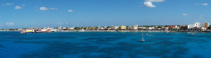 Wide panoramic photo of Cozumel town waterfront seen from the sea. Turquoise and deep-blue water in foreground, low colorful buildings and ferry terminal along the shore, a single small sailboat anchored, sunny blue sky.